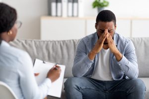 Desperate black man suffering from nevrous breakdown or depression, having session at psychologist's office, panorama. Stressed male patient meeting with therapist, receiving professional assistance